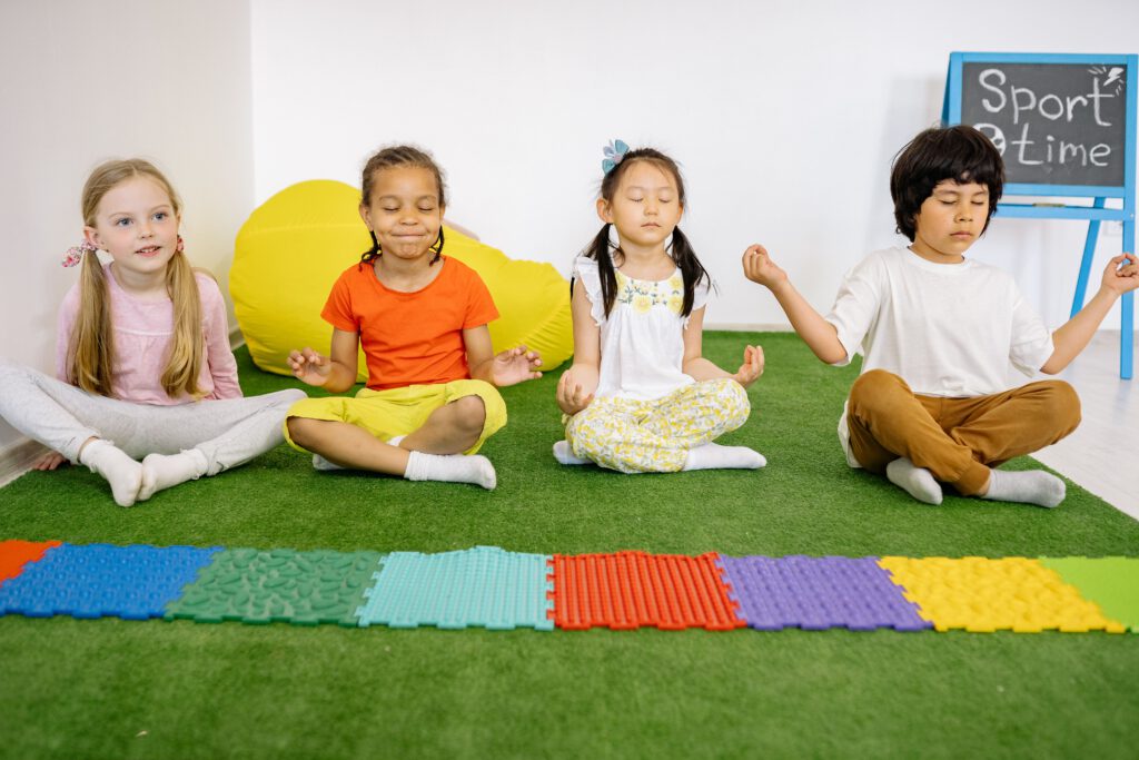 Kinder sitzen im Schneidersitz auf einer grünen Matte und halten die Hände vor der Brust.