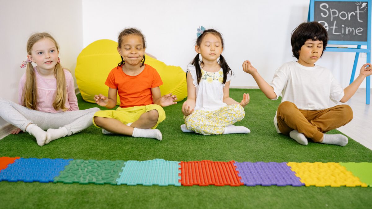 Zwei Kursleiterinnen sitzen mit einer Gruppe Vorschulkinder im Kreis auf Matten und üben gemeinsam eine ruhige Atemübung.