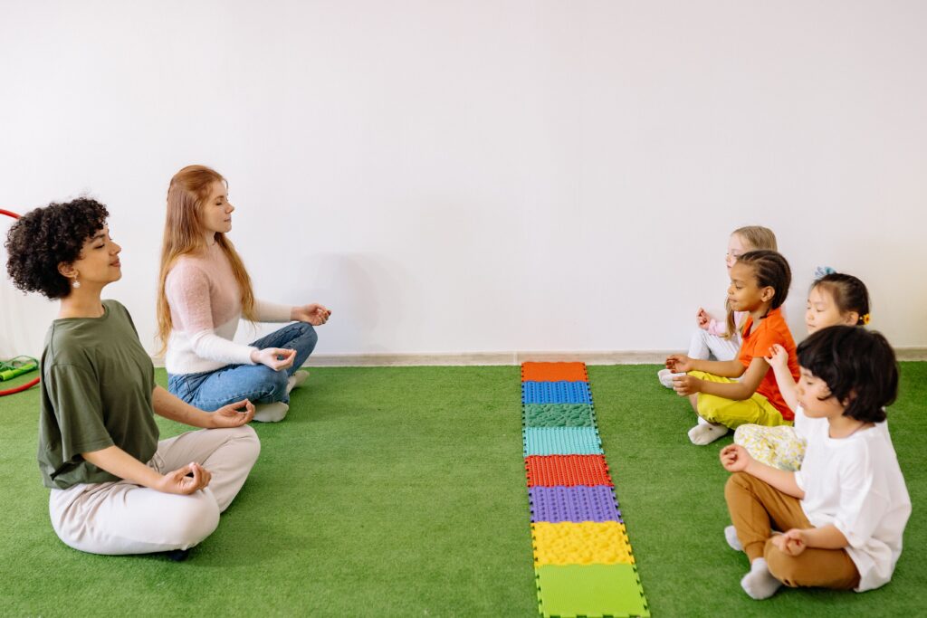 Zwei Kursleiterinnen sitzen mit einer Gruppe Vorschulkinder im Kreis auf Matten und üben gemeinsam eine ruhige Atemübung.